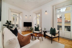 Un salón espacioso con un sofá blanco, una mesa de centro y dos sillones. Un televisor de pantalla plana está montado en la pared, y hay ventanas amplias con vistas a la ciudad.