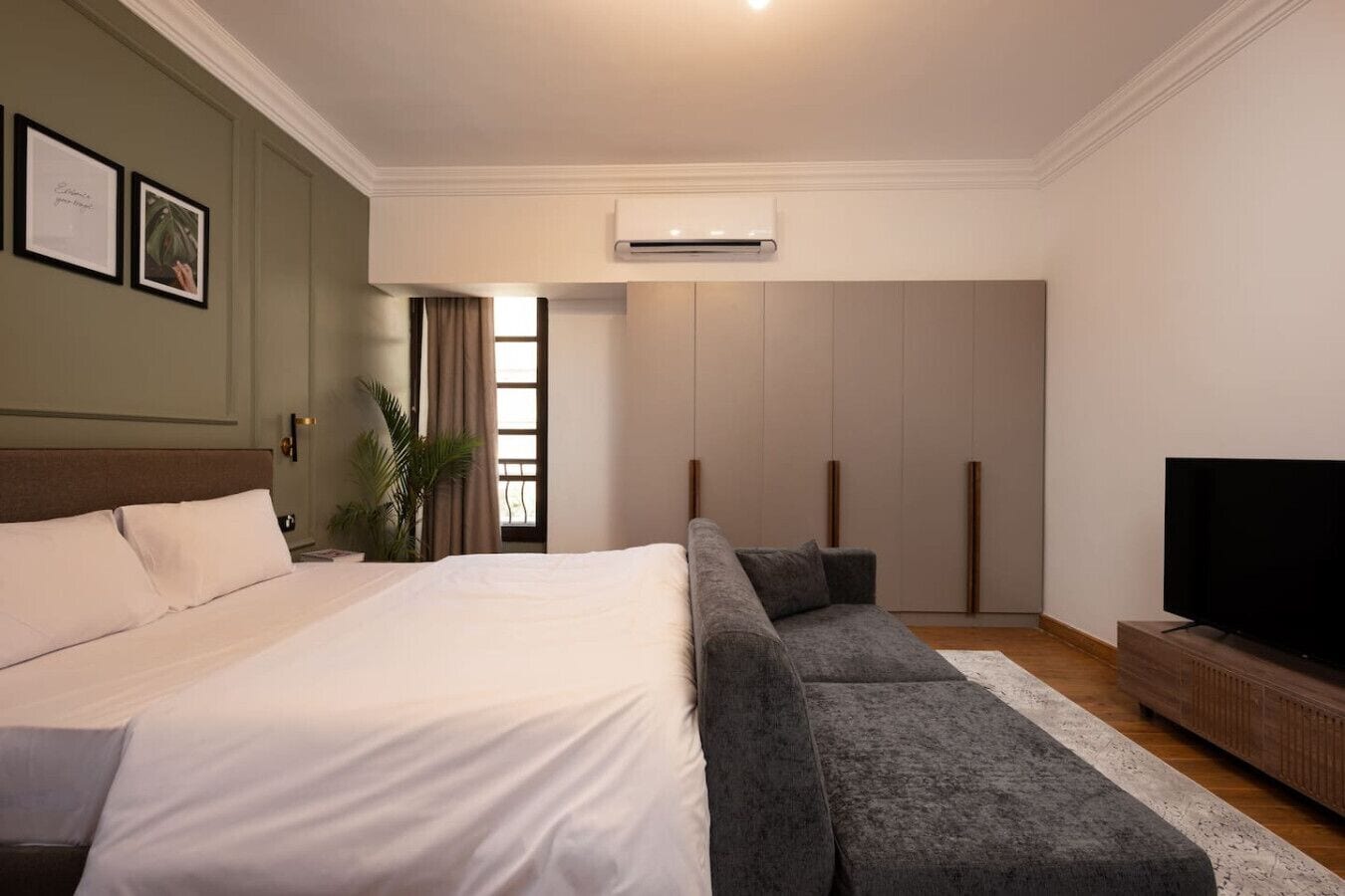 Une chambre moderne avec un grand lit, un canapé gris, une télévision et un appareil de climatisation.