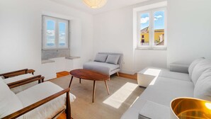 Ein modernes Wohnzimmer mit einer Couch, einem Holztisch und einem Essbereich mit Stühlen. Zwei Fenster bieten Ausblicke nach draußen.