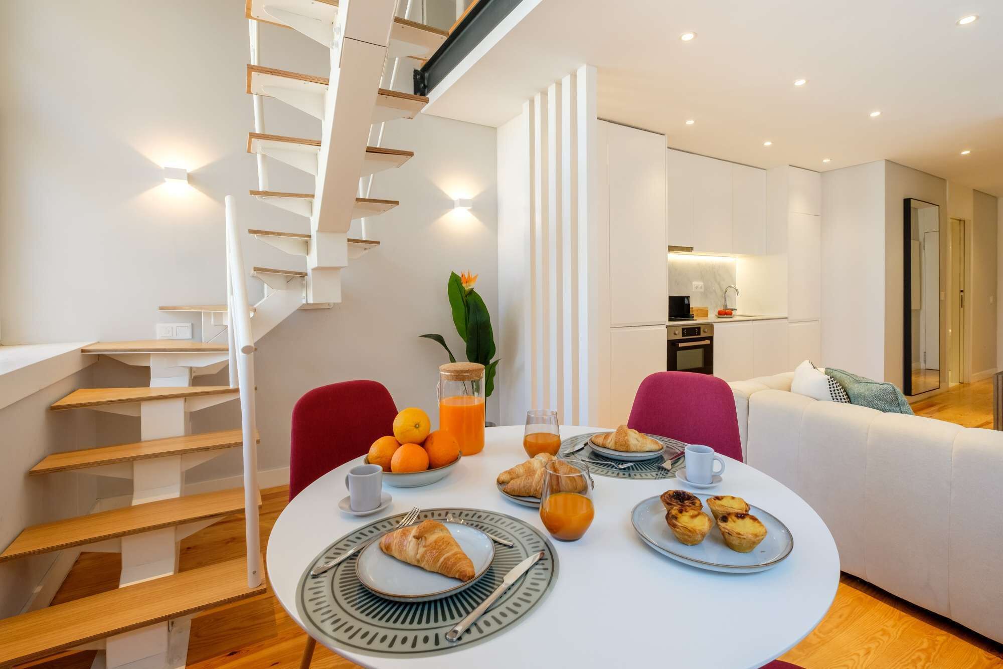 A modern dining area with a round table set for breakfast, a staircase, and a kitchen in the background.