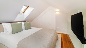 Ein Schlafzimmer mit schrägem Dach, einem Oberlicht, einem Bett mit weißen und grünen Kissen, einer beigen gesteppten Tagesdecke, Holzboden und einem an der Wand montierten schwarzen Fernseher.