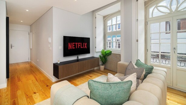 Ein modernes Wohnzimmer mit einem an der Wand befestigten Flachbildfernseher, einer Couch mit vielen Kissen und einem großen Fenster mit Ausblick.