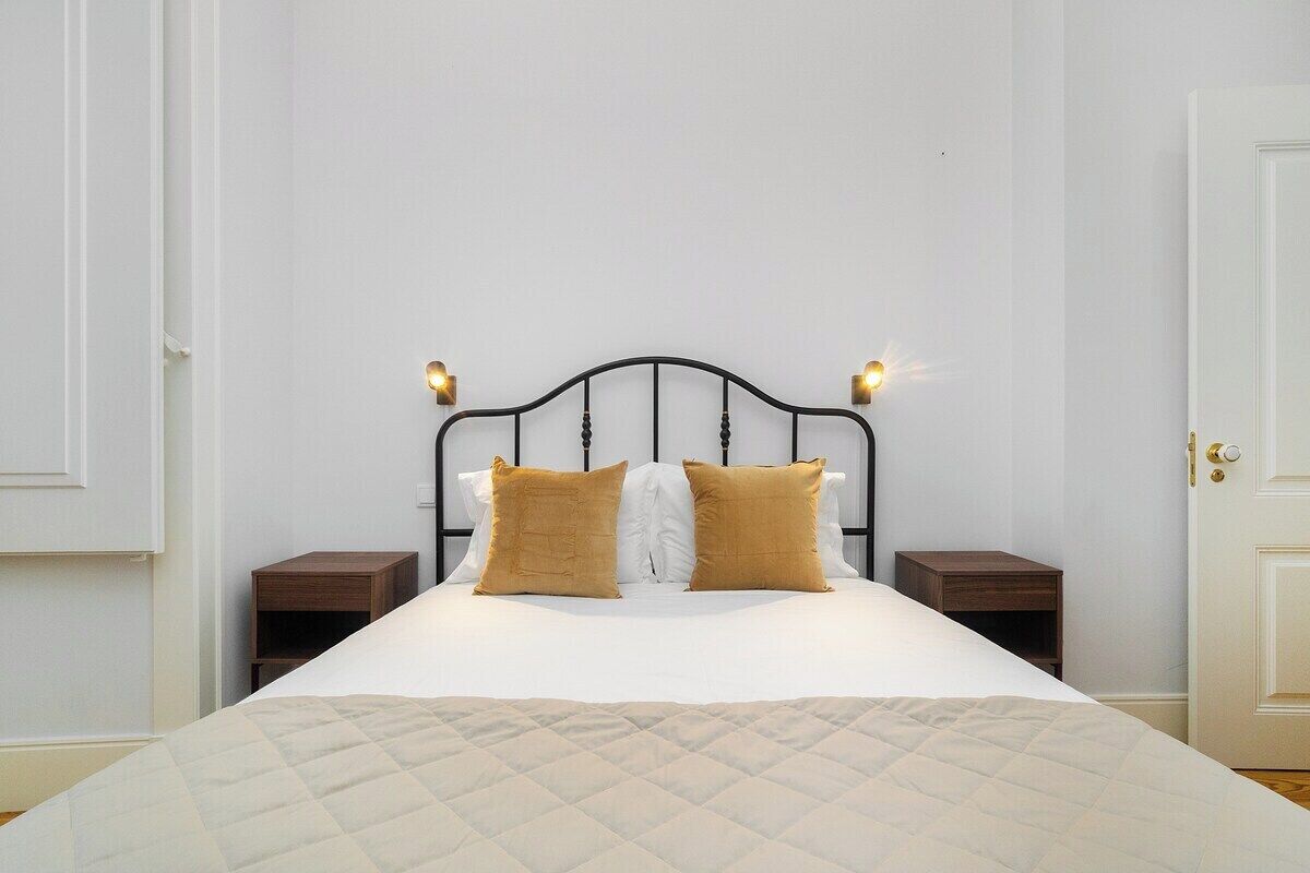 Una cama bien hecha con cubrecama acolchado, dos mesitas de noche con lámparas y una puerta a la derecha.