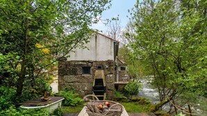 A rustic stone building with a wooden deck, surrounded by lush greenery and a river. - Riverfront Retreat with Exclusive Waterfall Views (Baião)