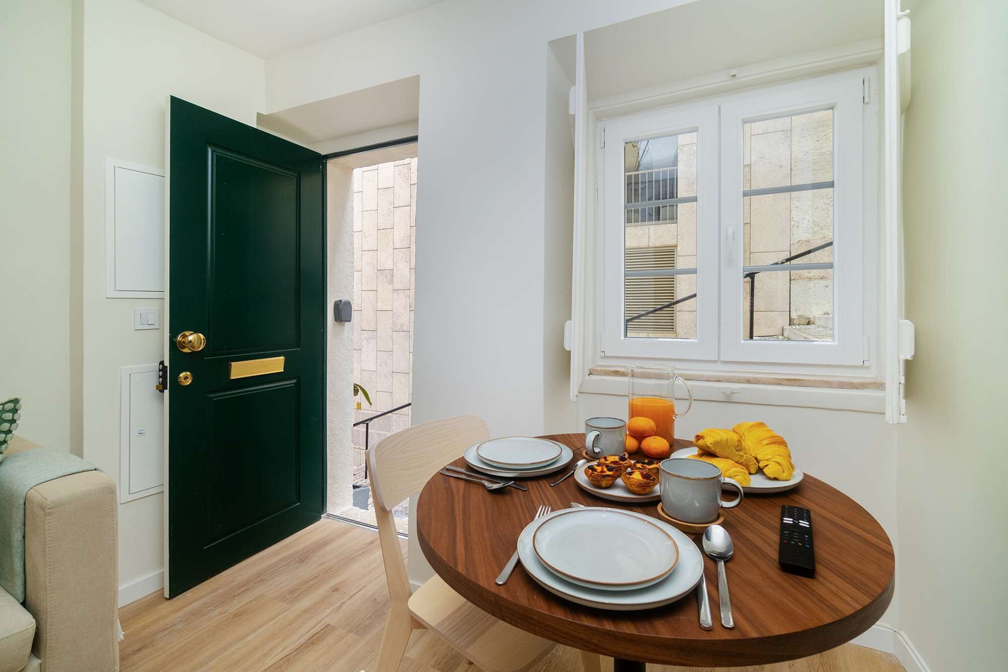 A dining area with a round wooden table set for breakfast, a green door, and a window with a view of a building outside.