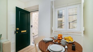 A dining area with a round wooden table set for breakfast, a green door, and a window with a view of a building outside. - Modern Flat in Bica’s Historic Charm (Lisbon)