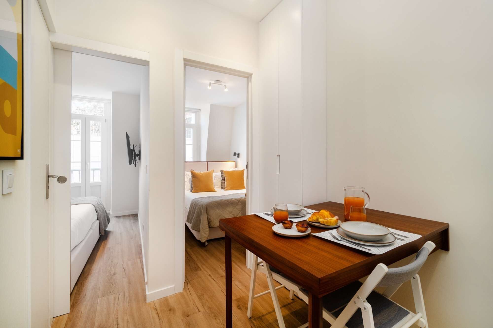 A compact room with a wooden dining table set for breakfast, a bed with a yellow pillow, and a white wardrobe.