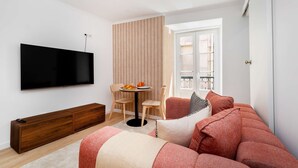 A modern living room with a flat-screen TV, a wooden bench, a round table with a chair, and a sofa with red cushions. - Charming House in Bica Neighbourhood (Lisbon)