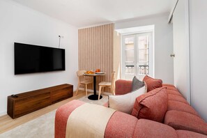 Een moderne woonkamer met een flatscreentelevisie, een houten bankje, een ronde tafel met een stoel en een bank met rode kussens.