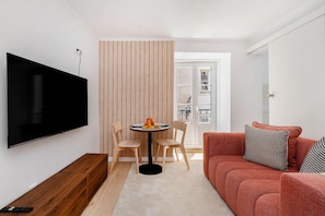 Een woonkamer met een flatscreen televisie, een ronde eettafel met stoelen, een rode bank met bedekte kussens en een houten scheidingswand.