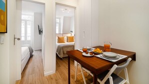 A compact room with a wooden dining table set for breakfast, a bed with a yellow pillow, and a white wardrobe. - Charming House in Bica Neighbourhood (Lisbon)