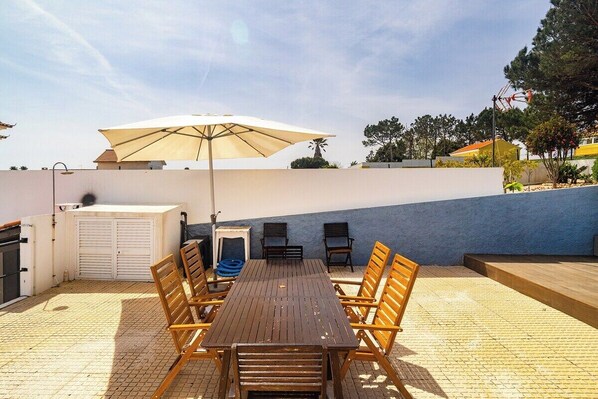 Une terrasse extérieure avec une table et des chaises en bois, un grand parasol et un tuyau d’arrosage bleu.