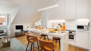 Une cuisine moderne avec des meubles blancs, un îlot central doté de tabourets, un téléviseur à écran plat et un coin repas comprenant un canapé et une table basse.