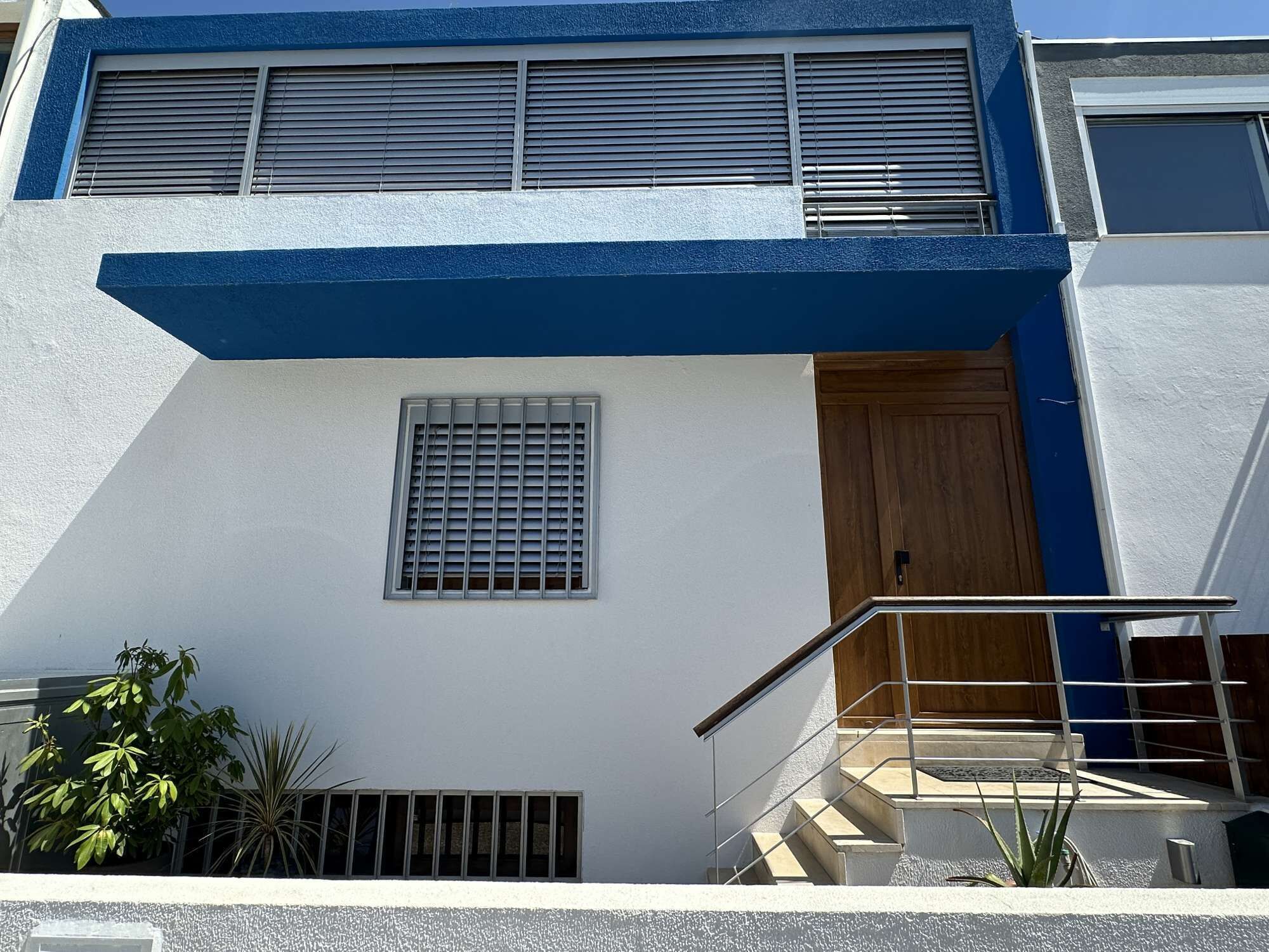 A modern two-story building with a blue and white facade, a wooden door, and a small window with a metal grill.