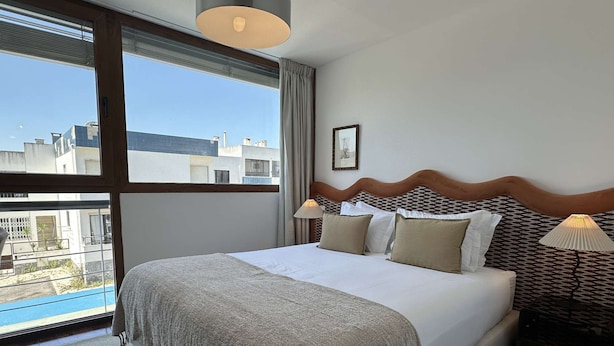 Ein ordentlich bezogenes Bett mit grauer Decke, zwei Nachttischlampen und Blick auf Gebäude durch ein großes Fenster.