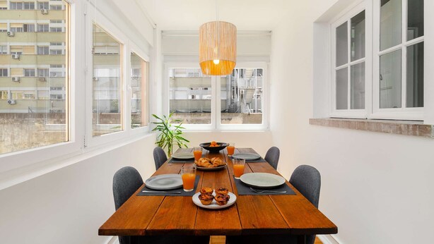 Comedor con mesa de madera, sillas, lámpara colgante y vista de un edificio a través de amplios ventanales.