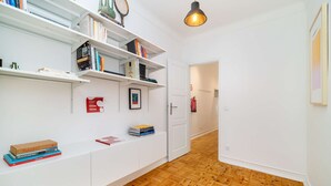 Un pasillo con paredes blancas, suelo de madera y estanterías blancas. Se ven libros, un cuadro enmarcado y un extintor.