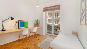 Un dormitorio moderno con suelo de madera, una cama, un escritorio con una silla, una lámpara de escritorio, un cuadro enmarcado, una planta en maceta y un balcón con vistas.