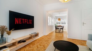 Una sala de estar moderna con una televisión de pantalla plana grande que muestra 'NETFLIX', suelo de madera, un reposapiés negro y un comedor con mesa y sillas.