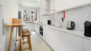 Una cocina moderna con armarios blancos, una mesa de comedor de madera con taburetes, una ventana con persianas y un extintor de incendios en la pared.