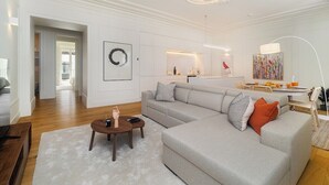 A modern living room with a grey sectional sofa, a wooden coffee table, and a white rug. There is a kitchen in the background with a dining area. - Timeless Design Meets Central Convenience (Lisbon)