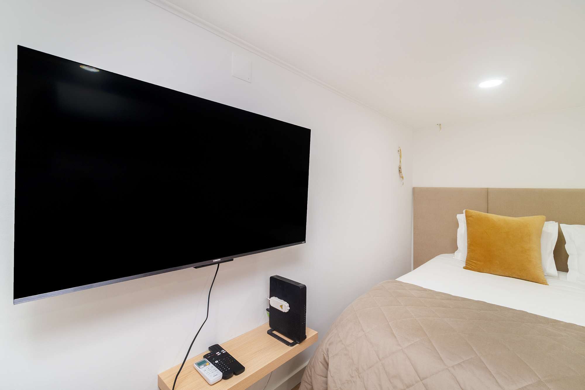 A hotel room with a large flat-screen TV mounted on the wall, a bedside table with a remote control, a black speaker, and a bed with a beige headboard and two pillows.