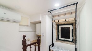 Een kleine kamer met een houten bankje, een raam met stenen omlijsting en een witte muur met een ingebouwde airconditioner.
