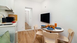 Une cuisine moderne avec des meubles blancs, un micro-ondes noir et une planche à découper en bois. Un coin repas avec une table ronde, des assiettes bleues et un téléviseur fixé au mur.