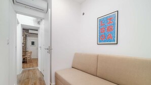 Un couloir avec un canapé beige, une œuvre d’art encadrée au mur et une porte donnant sur un espace repas.