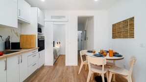 Eine kompakte Küche mit weißen Schränken, einer Holz Arbeitsplatte und einem eingebauten Backofen. Es gibt einen Essbereich mit einem runden Tisch und Stühlen, sowie eine kleine Frühstücksecke mit Orangensaft und Croissants.