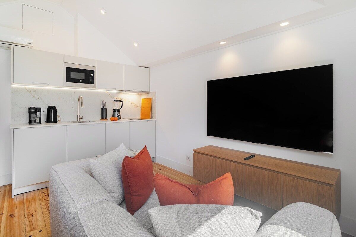 Een moderne woonruimte met een keukenhoek, een grijze bank met oranje kussens, en een grote flatscreen televisie aan de muur.