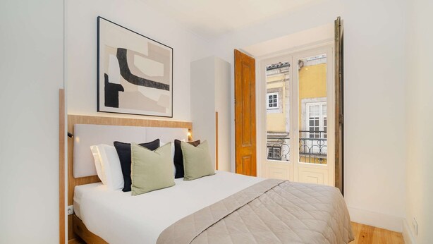 A bedroom with a bed, pillows, a headboard, a door, and a window with a view of buildings. - Live Like a Local in Vibrant Bairro Alto (Lisboa)