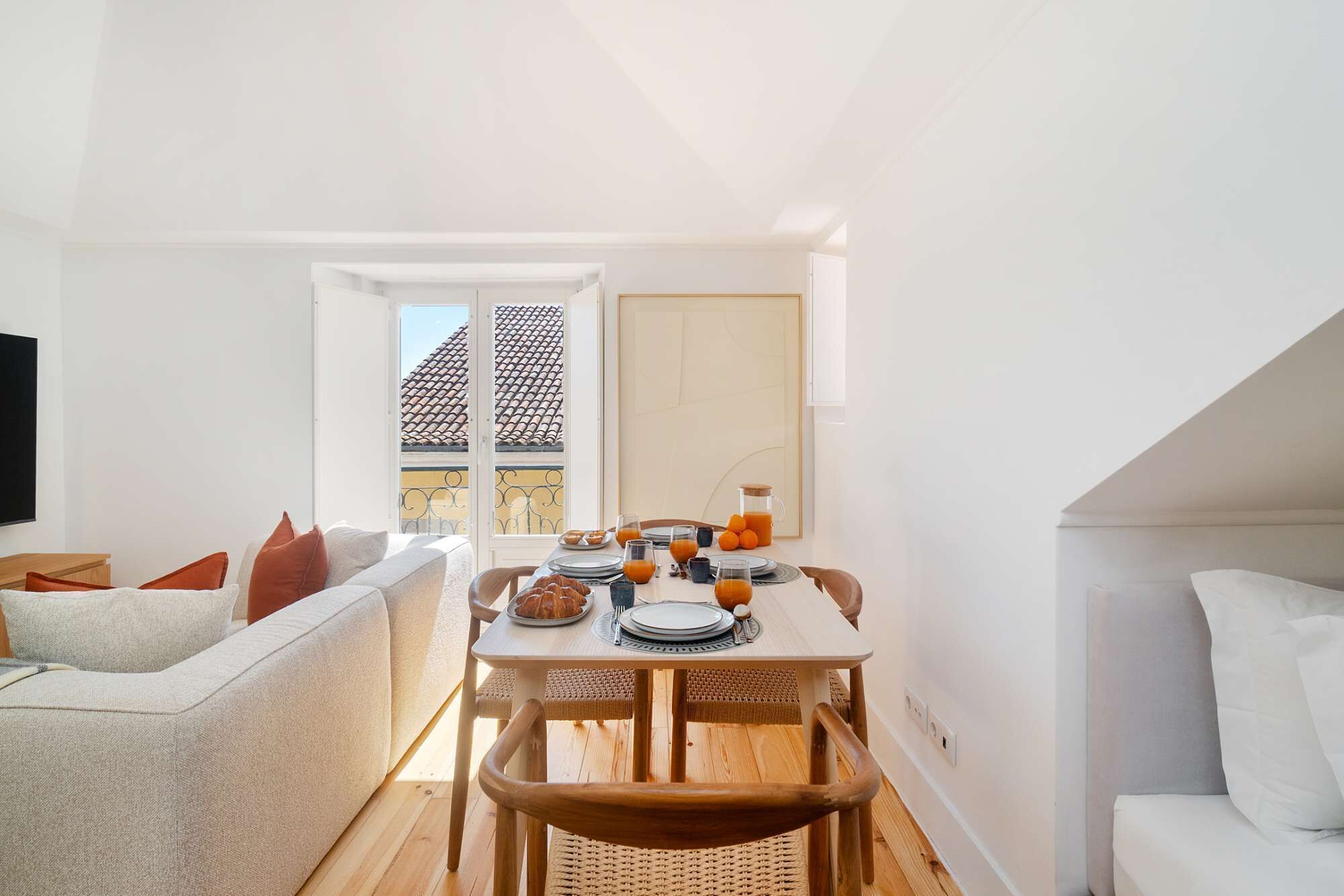 Uma sala de estar ampla e luminosa, com área de jantar, sofá e vista para um telhado cerâmico através de uma janela.