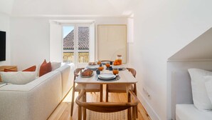 A bright, open-plan living space with a dining area, a sofa, and a view of a tiled roof through a window. - Live Like a Local in Vibrant Bairro Alto (Lisboa)