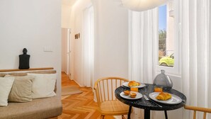 Ein modernes Hotelzimmer mit Essbereich, inklusive rundem Tisch mit Gebäckplatten und einem Glas Orangensaft, einem Holzsessel und Blick auf einen gelben Wagen draußen.