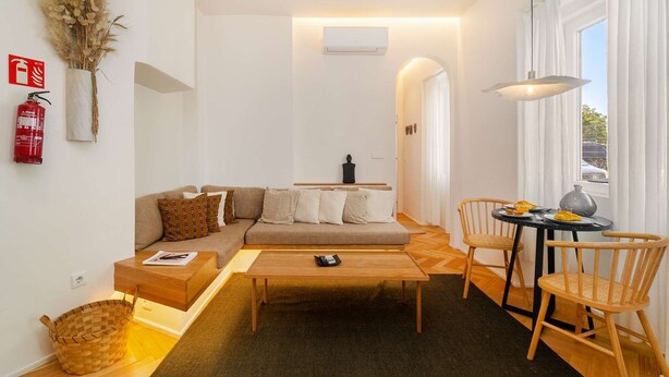 Ein modernes Wohnzimmer mit einer Couch, einem Couchtisch und Sesseln. An der Wand hängt ein Feuerlöscher, auf einem Tisch steht eine Vase mit Trockenpflanzen und es gibt ein Fenster mit Vorhängen.