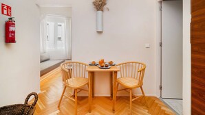 A room with a wooden floor, two wicker chairs, a small table with plates and glasses, a vase with dried flowers, a fire extinguisher, and a door. - Lisbon Charm: Walk to Graça Viewpoints (Lisbon)