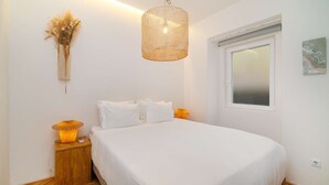 A bedroom with a large bed, a bedside table with a lamp, a wall-mounted light fixture, and a window with a view. - Lisbon Charm: Walk to Graça Viewpoints (Lisbon)