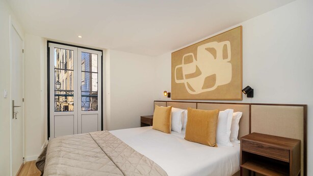 A hotel room with a large bed, a wooden nightstand, a wall-mounted lamp, and a window with a view of buildings. - Old Town Flat with a Modern Touch (Lisbon)