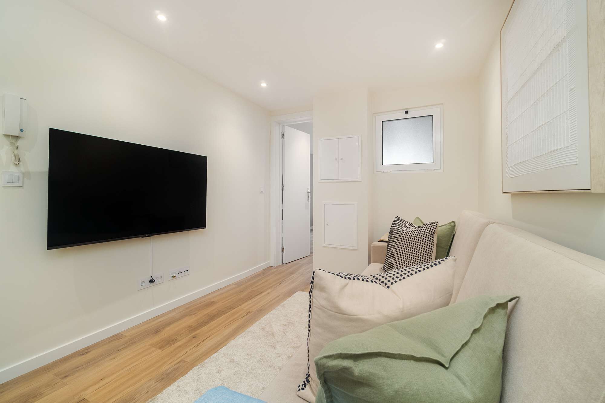Een moderne woonkamer met een flatscreen televisie aan de muur, een beige bank met decoratieve kussens, en een lichtkleurig vloerkleed.