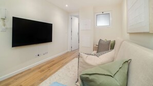 A modern living room with a flat-screen TV mounted on the wall, a beige sofa with patterned cushions, and a light-colored carpet. - Old Town Flat with a Modern Touch (Lisbon)