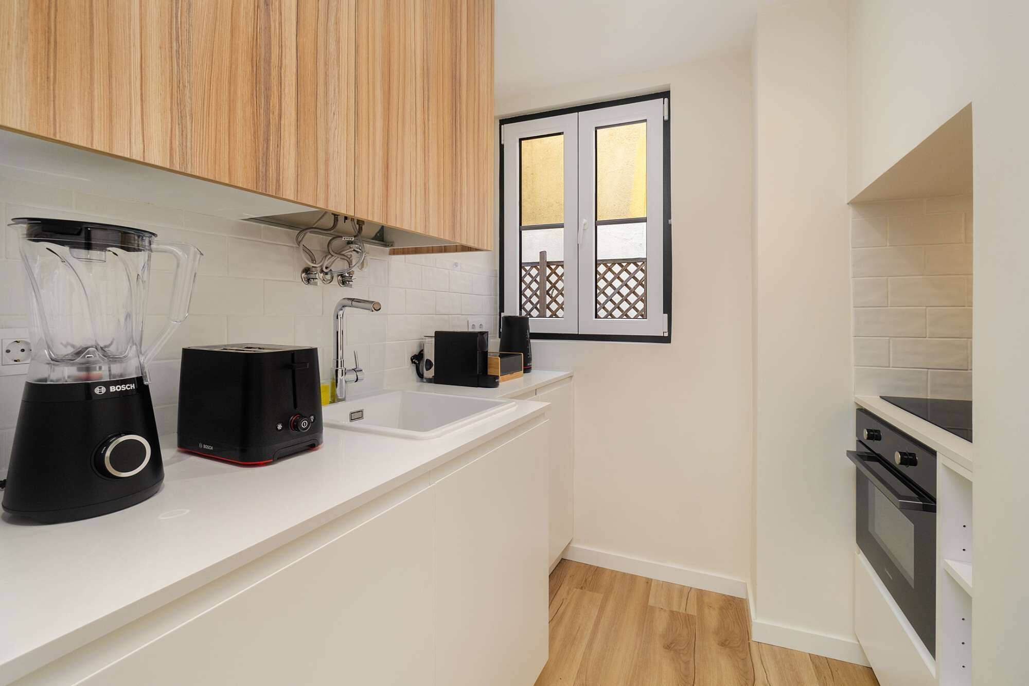 Una cocina compacta con encimeras blancas, una licuadora Bosch, un tostador y una ventana pequeña con vistas a una terraza de madera.