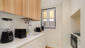 A compact kitchen with white countertops, a Bosch blender, a toaster, and a small window with a view of a wooden deck. - Old Town Flat with a Modern Touch (Lisbon)