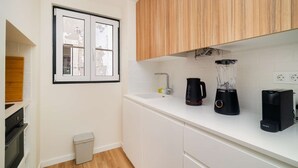 A kitchen with white cabinets, a Bosch blender, a coffee maker, and a window with a view of trees. - Old Town Flat with a Modern Touch (Lisbon)