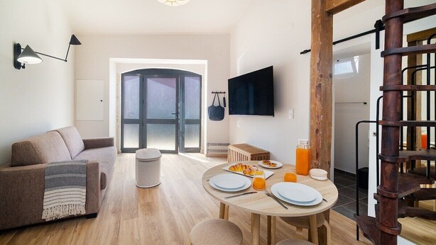 A spacious living area with a sofa, a dining table set with plates and a glass of orange juice, and a television mounted on the wall. - Sunny Studio Stay in Central Oeiras (Oeiras)