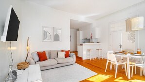 Una sala de estar moderna con un sofá, una mesa de comedor con sillas, una cocina al fondo y un televisor montado en la pared.