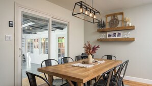 Un área de comedor con una mesa de madera, sillas negras, una lámpara colgante y un estante en la pared con adornos.