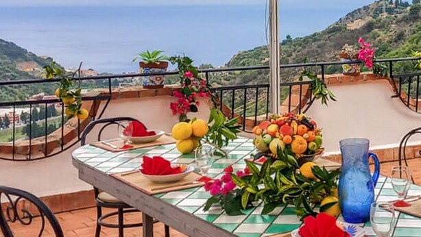 A table set for two with a view of the sea, a blue pitcher, and a bowl of fruit on a balcony overlooking a hilly landscape. - Nice home in Gioiosa Marea (Gioiosa Marea)