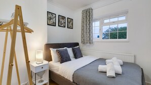A bedroom with a bed, a bedside table with a lamp, a window with a view, and a wooden easel. - Modern 4 Bedroom Hse Garden in Feltham (Feltham)