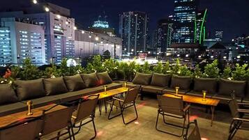 A rooftop lounge with seating and tables, overlooking a cityscape at night.